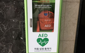 건물 내부 벽에 설치된 자동심장충격기(AED) 보관함. 빨간색 AED 장비가 투명문 안에 보관되어 있음